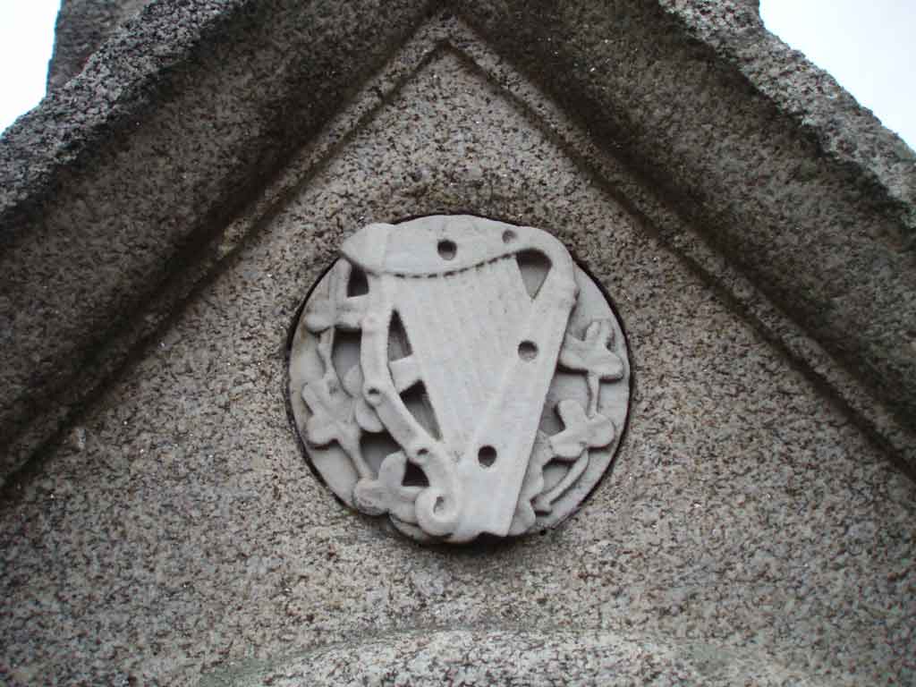 Graphite pencil sketch of a carved stone medallion depicting a harp surrounded by small clover motifs, set into a triangular pediment, grey stone tones