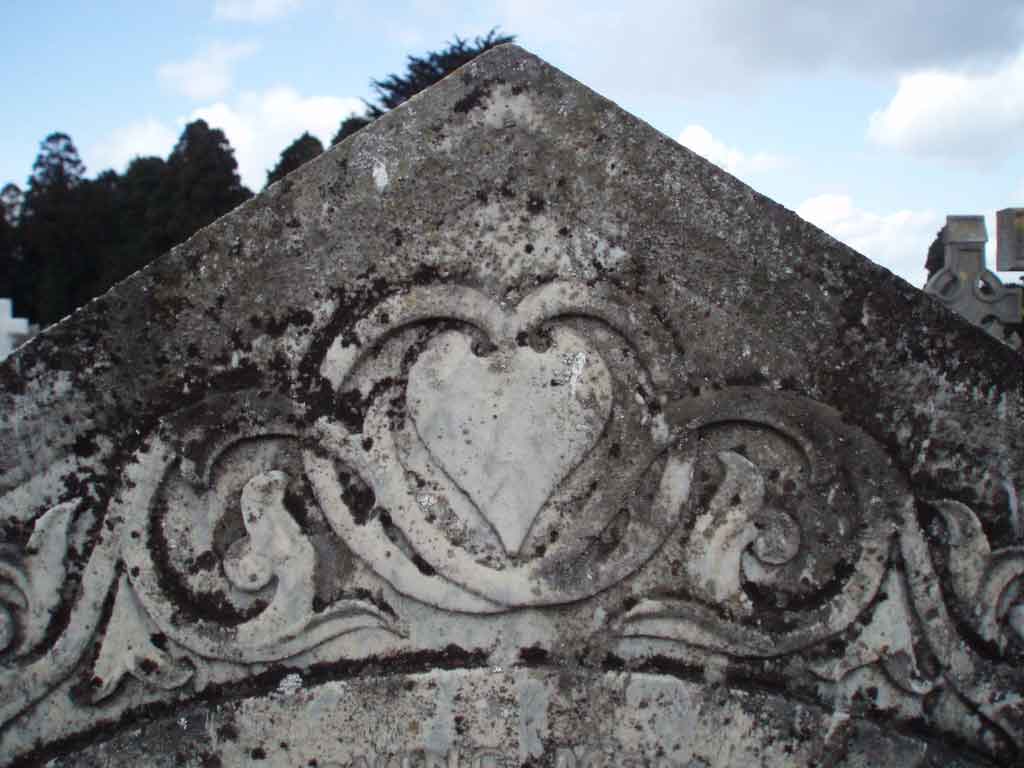 Graphite pencil sketch of a weathered gravestone corner with carved heart and scroll motif, grey and white stone tones, cloudy sky behind