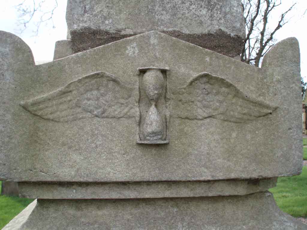 Graphite pencil sketch of a carved stone monument with a winged hourglass motif, bare tree branches and grass behind, sombre grey tones